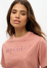 Camiseta de algodón rosa con un logo bordado de "Superdry" en la parte frontal. El diseño cuenta con un cuello redondo clásico y mangas cortas.