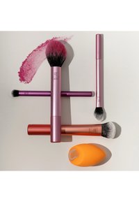 Cinq outils de maquillage disposés sur une surface claire : quatre pinceaux à manche rose avec de la poudre colorée et une éponge maquillage orange avec une tache de fond de teint.