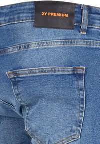 Jeans de mezclilla azul con un bolsillo trasero, costuras naranjas y una etiqueta de cuero oscuro con "2Y PREMIUM" grabado en naranja.