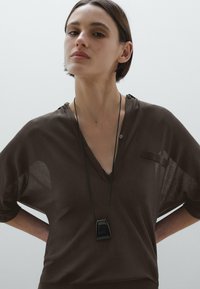 Brown, sheer, short-sleeve top with a collar and a button detail, paired with a black geometric pendant necklace. Smooth texture.