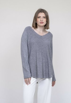 Femme debout, portant un pull ample gris à manches longues et un pantalon blanc, expression neutre, fond clair uni.