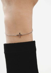 Pulsera de cadena de plata con un colgante de cruz horizontal. La pulsera se lleva en una muñeca, parcialmente cubierta por una manga negra.