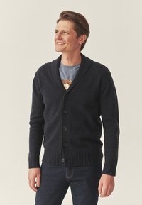 Tumši jūrā zila adīta džemperis ar šalli, ar pieciem pogām, ribotām piedurknēm un apakšmalu, uzvilkts virs gaiši zilas t-krekla.