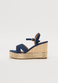 Marineblå kilehæl sandal med kryssende stropper, en teksturert plattform av naturlig fiber og en gullmetallisk detalj på bunnen.
