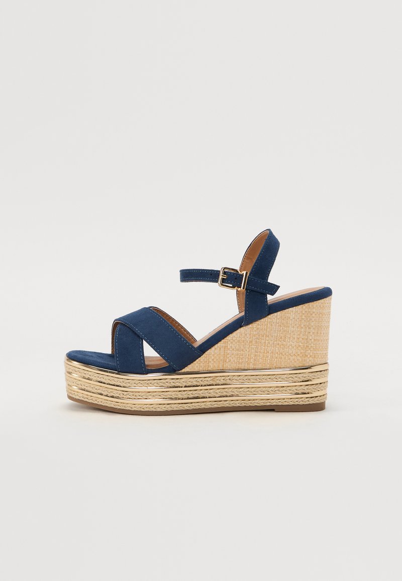 Marineblå kilehæl sandal med kryssende stropper, en teksturert plattform av naturlig fiber og en gullmetallisk detalj på bunnen.