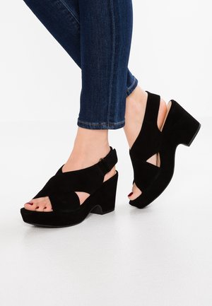 Voeten in zwarte suède plateauzandalen met gekruiste bandjes, gedragen met donkerblauwe skinny jeans, op een witte vloerachtergrond.