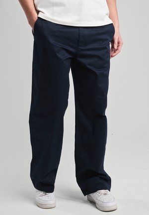Person trägt lockere dunkelblaue Hose und weiße Turnschuhe, mit teilweise in die Taschen gesteckten Händen, stehend vor einem einfarbigen hellen Hintergrund.