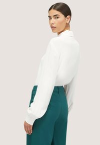 Camicia bianca a maniche lunghe con colletto, abbinata a pantaloni verdi a vita alta. La camicia ha un tessuto leggermente texturizzato.