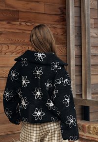 Sort fleecejakke med hvid blomstret broderi, der har en bred krave og en afslappet pasform. Tekstureret, hyggeligt stof med løse ærmer.