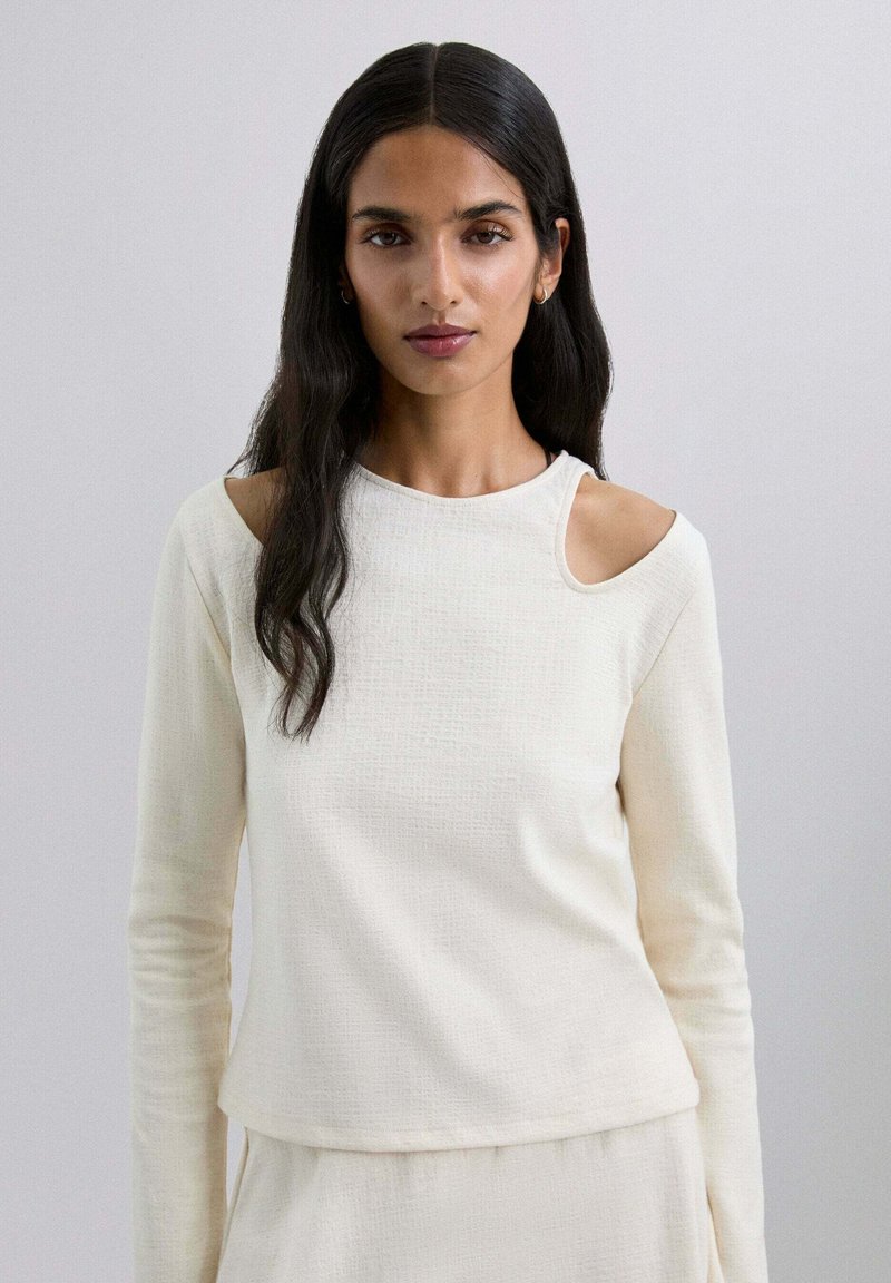 Haut à manches longues en crème clair, en tissu texturé. Comprend un détail d'épaule ouverte et un col arrondi. Design simple et ajusté.