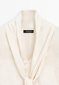 Gros plan d'un chemisier texturé couleur crème avec un col drapé et un détail noué à l'avant, avec une étiquette Massimo Dutti.