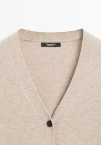 Beige kardigan med V-hals och en mörk knapp. Tillverkad av mjukt tyg, med en slät textur och minimalistisk design. Märkesetikett närvarande.
