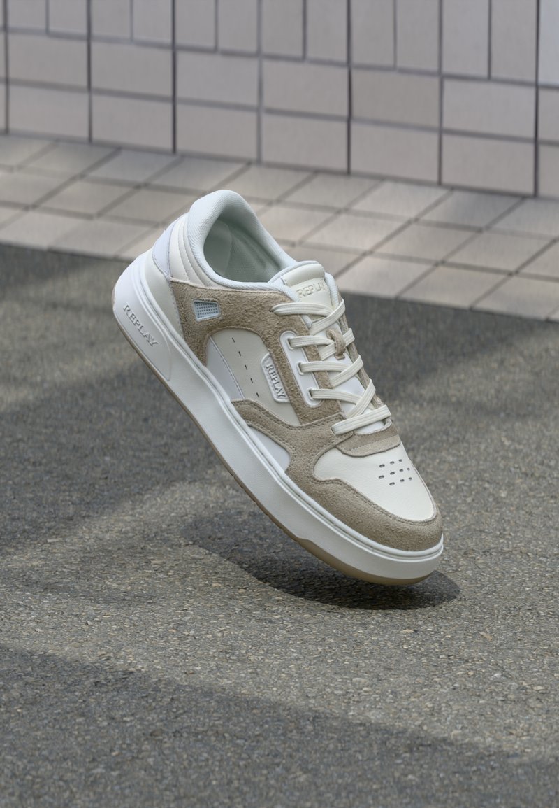Baskets Replay blanches et beige avec lacets, panneaux en daim et semelle en caoutchouc flottant au-dessus d'un pavé en asphalt près d'un mur carrelé.