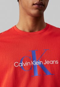 Camiseta de algodón roja con un audaz logotipo gráfico azul "CK" y "Calvin Klein Jeans" en blanco, cuello redondo, mangas cortas, textura suave.