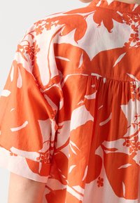Vue de dos d'un chemisier orange et blanc à grand imprimé floral et manches courtes, fabriqué en tissu léger.