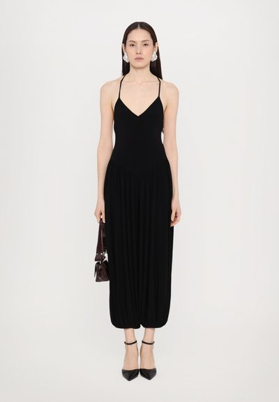 Schwarzer ärmelloser Jumpsuit mit V-Ausschnitt, gerafftem Bund und schmal zulaufenden Beinen. Kombiniert mit schwarzen spitzen Absätzen und einer kleinen burgunderfarbigen Tasche.