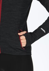 Black athletic jacket with red accents, featuring zippered pockets and thumbholes, made from a textured, moisture-wicking fabric.