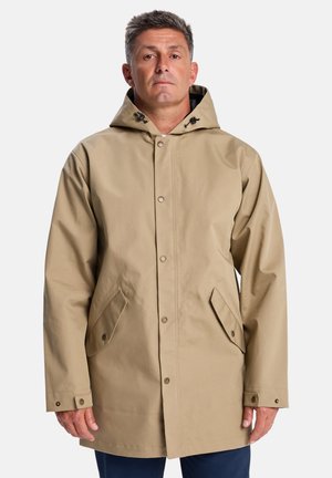 Een man staat met een lichtbruin regenjack met capuchon, voorzien van zijzakken, tegen een effen witte achtergrond.