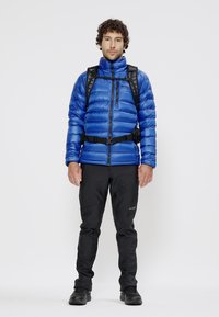 Doudoune bleue avec fermeture éclair à l'avant, portée sur une couche de base noire ; pantalon softshell noir ; comprend un sac à dos noir et des accents de ceinture.