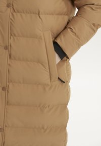 Tan pufferjakke med quiltet design, der har knaplukning foran og sidelommer. Glat stof med en let skinnende overflade.