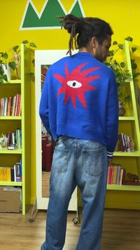 Niebieski sweter dziergany z czerwonym, graficznym motywem oka na plecach. Połączony z jasnoniebieskimi dżinsami. Charakterystyczna ściągaczowa krawędź i mankiety.