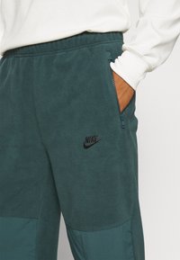 Person iført mørkegrønne Nike fleecebukser med lynlåslomme, hånd i lommen og en hvid langærmet top.