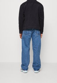 Černá kordová bunda přes modrý denimový outfit, který zahrnuje volné džíny a vzorované tenisky. Kontrast textur mezi materiály.