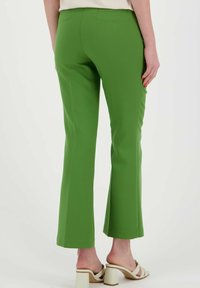 Pantalons évasés verts en tissu lisse, avec une taille mi-haute et une coupe épurée. Portés avec des sandales à talons carrés beiges.