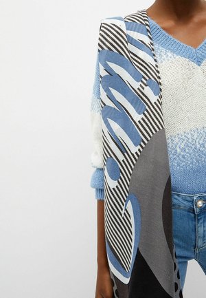 Personne portant un pull en tricot bleu et blanc et un jean, tenant une grande écharpe aux motifs abstraits noirs, blancs, bleus et gris sur une épaule.