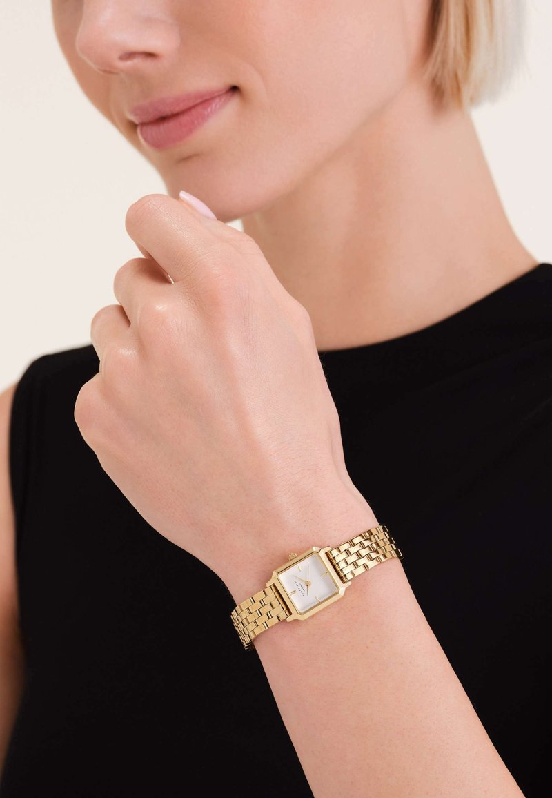 Montre dorée avec un cadran carré blanc, design de bracelet en maillons, et une petite couronne sur le côté, portée sur une main contre un haut noir.