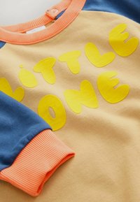 Light beige sweatshirt with yellow "LITTLE ONE" text, blue sleeves, and an orange ribbed collar and cuff. Soft cotton fabric.