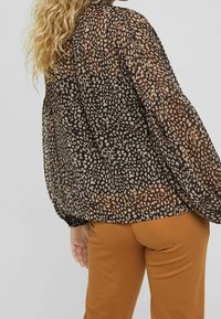 Blusa nera trasparente con un motivo maculato in marrone e crema. Maniche lunghe con polsini arricciati. Abbinata a pantaloni aderenti marroni.