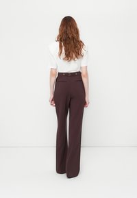 Une personne avec de longs cheveux ondulés porte un haut blanc à manches courtes rentré dans un pantalon large taille haute, marron foncé, faisant face à un mur blanc uni.