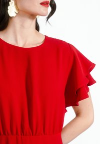 Rochie roșie cu mâneci scurte, volanate; material moale, ușor; decolteu rotund; talie elastică; asociată cu cercei mari în formă de scoică.