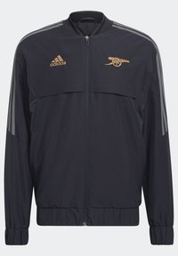 Veste noire Adidas à fermeture éclair avec des bandes grises sur les manches, logo Adidas doré sur la poitrine gauche et emblème canon doré sur la poitrine droite.
