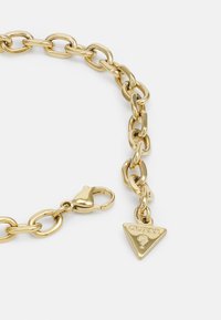 Pulsera de cadena de metal dorado con eslabones ovalados, un cierre de langosta y un charm triangular con "GUESS" grabado en él.