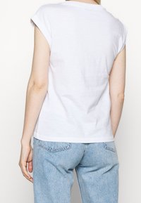Persona con una camiseta blanca de manga corta y jeans de mezclilla azul claro, mostrada de espaldas contra un fondo blanco.