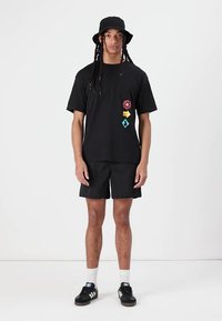Must musta säravate graafikaga, millele on lisatud mustad lühikesed püksid. Aksessuaarideks on must bucket hat ja mustad tennisetossud valgete detailidega.