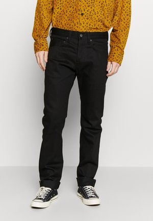 Personne portant un jean noir droit, une chemise jaune moutarde à manches longues avec des motifs noirs, et des baskets noires et blanches.
