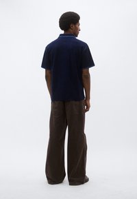 Σκούρο μπλε πουκάμισο με κοντά μανίκια, υφαντό ύφασμα, γιακάς και μπλε λεπτομέρειες. Συνδυασμένο με φαρδιά καφέ παντελόνια, θέα από πίσω.