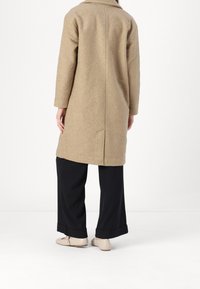 Béžový vlněný kabát s jemnou texturou, s švem na zádech a klopovým límcem. Kombinován s černými kalhotami s širokými nohavicemi a světlými botami.