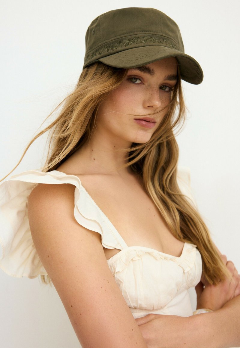 Jeune femme aux longs cheveux blonds porte une casquette vert olive et un haut blanc sans manches à volants sur un fond clair uni.