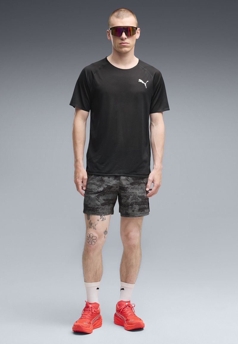 Άνδρας φοράει μαύρο t-shirt Puma, σκούρο σορτς με σχέδιο, κόκκινα αθλητικά παπούτσια Puma, λευκές κάλτσες και ανακλαστικά αθλητικά γυαλιά ηλίου, και στέκεται στραμμένος προς τα εμπρός.
