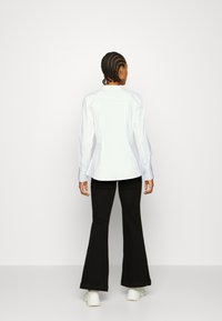 Camisa blanca de manga larga con un diseño ajustado, combinada con pantalones de campana negros y zapatillas blancas. Tejido suave, cuello clásico y detalles de botones.