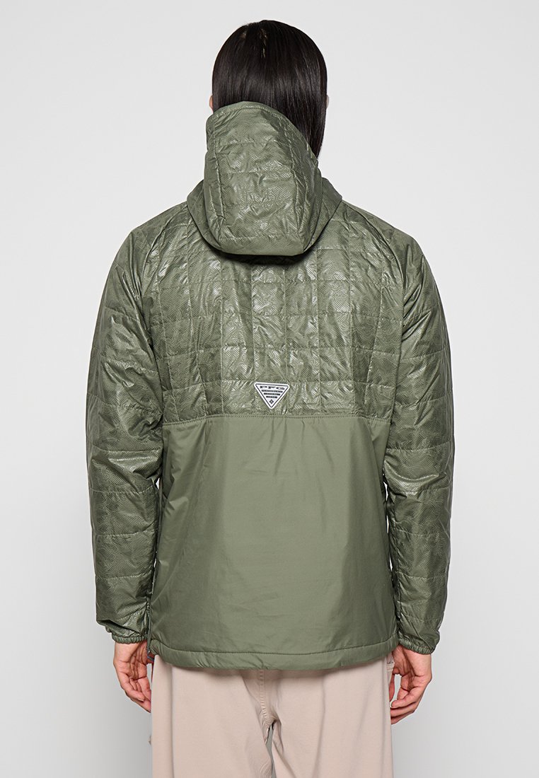 Person mit dunklem Haar, der eine zweifarbige, olivgrüne, gesteppte Jacke mit Kapuze trägt, von hinten vor einem schlichten Hintergrund betrachtet.