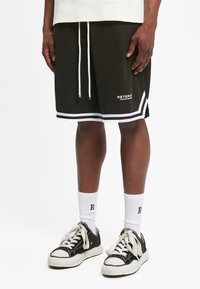 Svart mesh idrettsshorts med hvite detaljer, snøring i midjen og logobolk. Kombinert med svarte joggesko og hvite sokker.