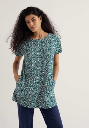 Kobieta z długimi kręconymi włosami, ubrana w turkusową tunikę w kwiaty z krótkim rękawem i granatowe spodnie, stoi z rękami w kieszeniach na jednolitym tle.