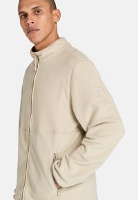 Beige fleecejacka med hög krage, framzipper och zippered sidofickor. Har en logotypapplikation på vänster ärm. Mjuk textur.