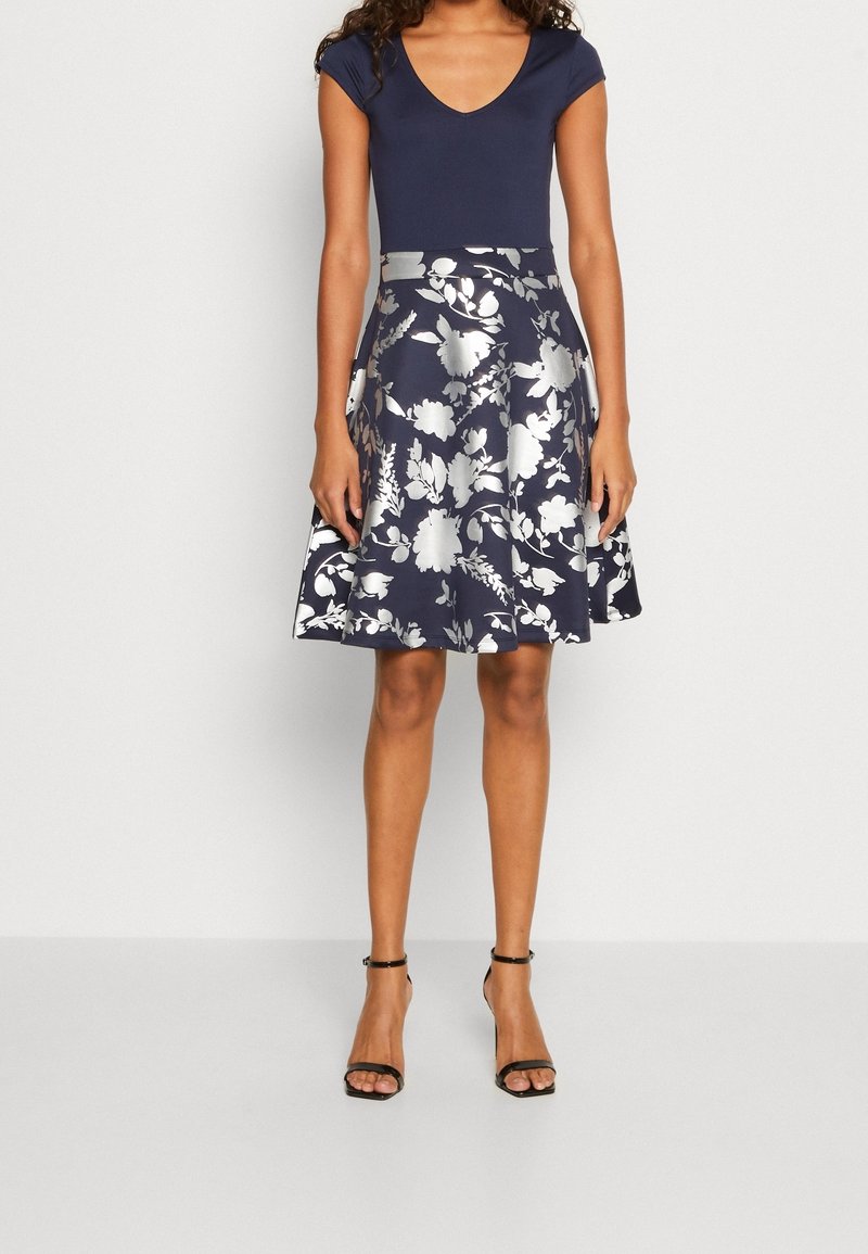 Femme portant un haut bleu marine à manches courtes et une jupe bleu marine longueur genou avec de grands motifs floraux blancs et des talons noirs à brides.