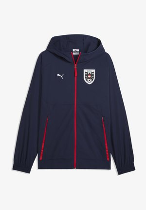 Marineblaue Kapuzenjacke mit rotem Reißverschluss und Taschenumschlägen, Puma-Logo auf der rechten Brust und schwarz-weiß-rotem Wappen mit Fußb all auf der linken Brust.